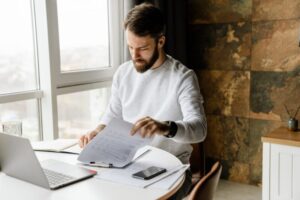 Bearded man working with laptop and papers while sitting at table