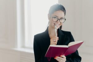 Glad successful budget analyst notes some text in notebook, smiles gladfully, holds pen