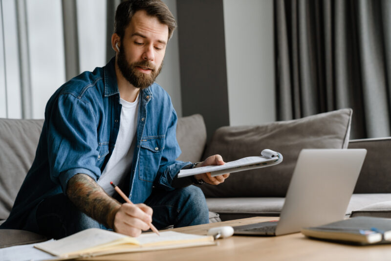 Bearded man writing down notes while making conference call on laptop