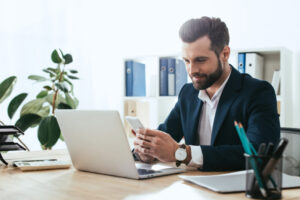 smiling and handsome advisor in suit using smartphone at workspace