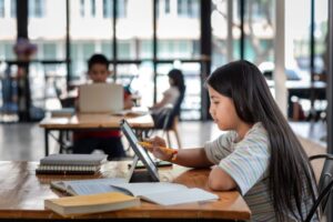 Side view of a girl holding a pen pointing at the tablet screen The book is placed on the table the