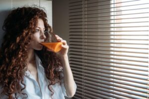 Woman standing by a windows blind drinking orange juice