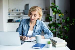 young blonde woman studying online and making notes