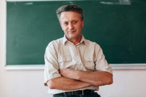 Happy teacher sitting folded a hands next a blackboard.- Image