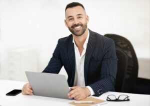 Glad confident caucasian mature man with beard in suit sits at table with laptop, has meeting
