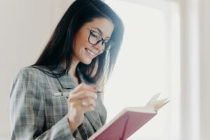 Woman makes notes in planner, does remote job, wearing glasses and formal clothes, poses near window