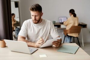 Young serious man making notes in copybook and looking at laptop screen
