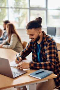 College student writing notes while e-learning on laptop in the classroom.