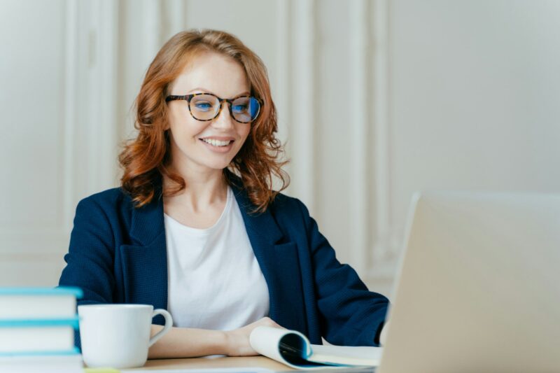 Glad lovely woman has ginger hair, positive smile, sits with laptop computer at desktop