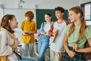 Positive teenage pupils with devices and backpacks talking while standing in classroom in school