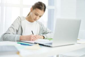 Concentrated latin teenage girl making notes in her notepad, using laptop while having online lesson