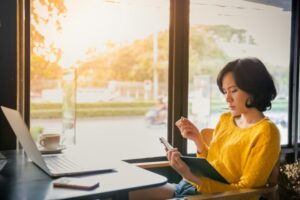 Young business woman in yellow dress sitting at table in cafe and writing in notebook.