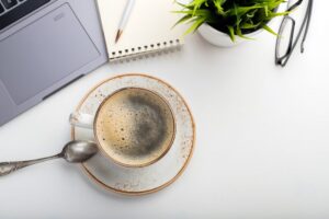 Desk with laptop, eye glasses, notepad, pen and a cup of coffee on a white table.