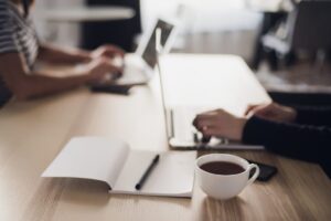 Close up shot of a cup of coffee, notebook with pencil or pen and hands typing on keyboard of a